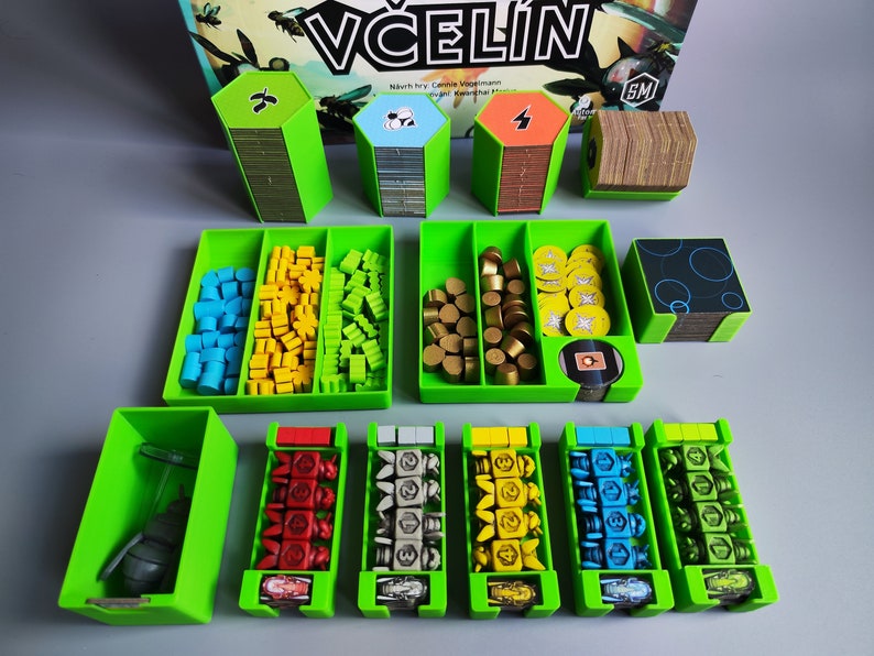 K&ouml;nnte beinhalten: Gr&uuml;ne Aufbewahrungsboxen f&uuml;r das Brettspiel "Včel&iacute;n" von Game Factory. Die Boxen enthalten verschiedene Spielsteine, darunter Holzw&uuml;rfel, Spielmarken und Karten. Der Spieltitel ist auf dem Karton sichtbar.