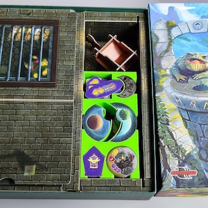 Può includere: Una scatola di gioco da tavolo verde e viola con un inserto verde e viola per riporre i pezzi del gioco. Il coperchio della scatola presenta un'illustrazione di una creatura verde simile a una rana con una corona in testa. Il testo sulla scatola recita "Kroni Av", "Norddeutsch" e "Ror", che probabilmente sono il titolo del gioco, la regione e l'editore.