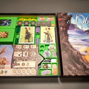 May include: An open box game with green and brown compartments. The game board is visible with a brown and white design. The box lid is closed and shows a colourful illustration of a landscape with a large tree and a yellow flower. The text "Obleva" is printed on the box lid.