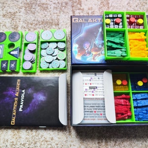 May include: A green and black board game box with the title "GALAKTICKÉ ALIANCE" and "PRAVIDLA" on the front. The box is open and shows the game components, including green and yellow plastic pieces, red and blue plastic pieces, and a rulebook.