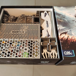May include: A gray and black board game box with a custom insert for organizing game pieces. The insert is made by "Inserty.cz" and features a honeycomb design. The box lid features a dramatic image of a battle scene with a large ship in the background. The text on the box lid reads "The Guild" and "Desk Top Game".