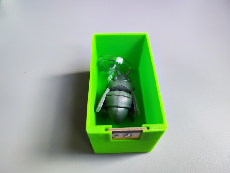 K&ouml;nnte beinhalten: Eine gr&uuml;ne Plastikbox mit einer grauen Insektenfigur im Inneren. Die Box hat die Nummer "3" auf der Vorderseite.