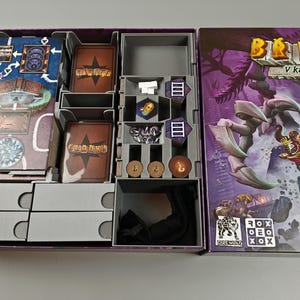 May include: Open board game box with various components. Includes game cards, tokens, and a game board with a map design. The box features the game title "Briniv V katakombách" with fantasy artwork.