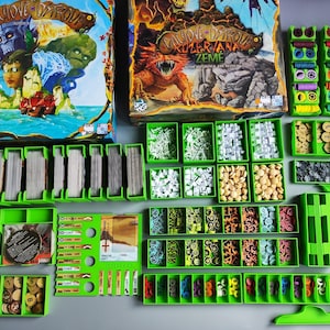 Può includere: Componenti di un gioco da tavolo disposti su una superficie grigia. Sono visibili due scatole di gioco, insieme a vari vassoi verdi pieni di carte, gettoni e pezzi del gioco. Le scatole presentano illustrazioni colorate di personaggi e creature fantasy.