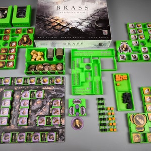Puede incluir: Bandejas organizadoras de plástico verde para el juego de mesa Brass: Birmingham. Las bandejas están etiquetadas con los nombres de los diferentes componentes del juego, como "Carbón", "Hierro" y "Textil".