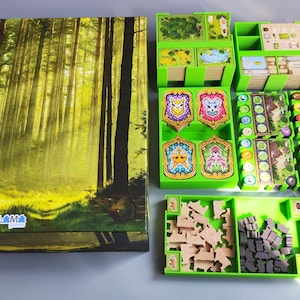 May include: Board game box with a forest scene and the text "Černý les". The game includes green storage trays, wooden game pieces, and colorful game cards. The box and components are arranged on a light surface.