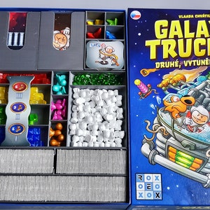 Może przedstawiać: Niebieskie pudełko z wkładką do gry planszowej Galaxy Trucker. Pokrywa pudełka przedstawia kolorową ilustrację statku kosmicznego z kreskówkowym astronautą i stworzeniem przypominającym ptaka. Tekst na pudełku brzmi "Galaxy Trucker" i "Druhé, Vytuněné Vydání".