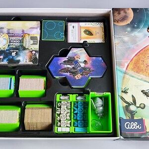 K&ouml;nnte beinhalten: Ein gr&uuml;ner und schwarzer Spielebox-Einsatz mit F&auml;chern f&uuml;r Karten, W&uuml;rfel und ein Miniatur-Raumschiff. Der Deckel der Box zeigt eine farbenfrohe Illustration eines Planeten und eines Raumschiffs.