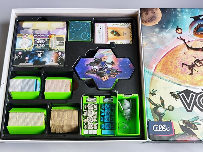 K&ouml;nnte beinhalten: Ein gr&uuml;ner und schwarzer Spielebox-Einsatz mit F&auml;chern f&uuml;r Karten, W&uuml;rfel und ein Miniatur-Raumschiff. Der Deckel der Box zeigt eine farbenfrohe Illustration eines Planeten und eines Raumschiffs.