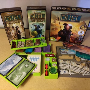 Peut inclure: Trois boîtes de jeu pour le jeu de société 7 Merveilles Duel, avec les extensions Agora, Panthéon et le jeu de base. Les boîtes sont sur une table avec des organisateurs de jeu verts et gris.