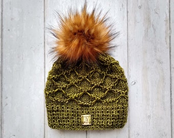 May include: A green knitted beanie with a brown faux fur pom pom. The beanie has a diamond pattern and a small gold tag.