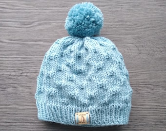 May include: A light blue knitted hat with a pom-pom on top and a small brown leather tag with the word "SANSAB" and a penguin logo.