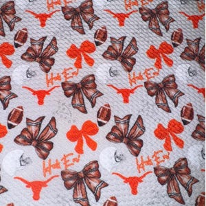 May include: A light gray fabric with a repeating pattern of brown bows, orange bows, footballs, and the text "Hook Em". The design also includes orange longhorn symbols, creating a collegiate theme.