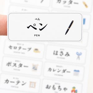May include: A rectangular white sticker featuring the Japanese hiragana characters for pen, the katakana characters for pen, the English word PEN, and a small illustration of a black pen with gold accents, part of a larger sheet of vocabulary labels.