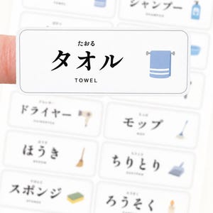 May include: A rectangular white adhesive label featuring the Japanese text タオル, the hiragana たおる, and the English word TOWEL in black, accompanied by a blue towel icon. The label is part of a larger sheet of similar vocabulary stickers.
