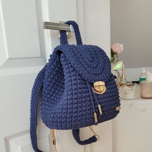 May include: A navy blue crocheted backpack with a gold chain strap and a gold clasp. The backpack is hanging on a white door.