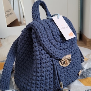 May include: A navy blue crocheted backpack with a gold metal clasp and a small tag that says "by Anna Nadel." The backpack is sitting on a patterned fabric with yellow, grey, and white triangles.