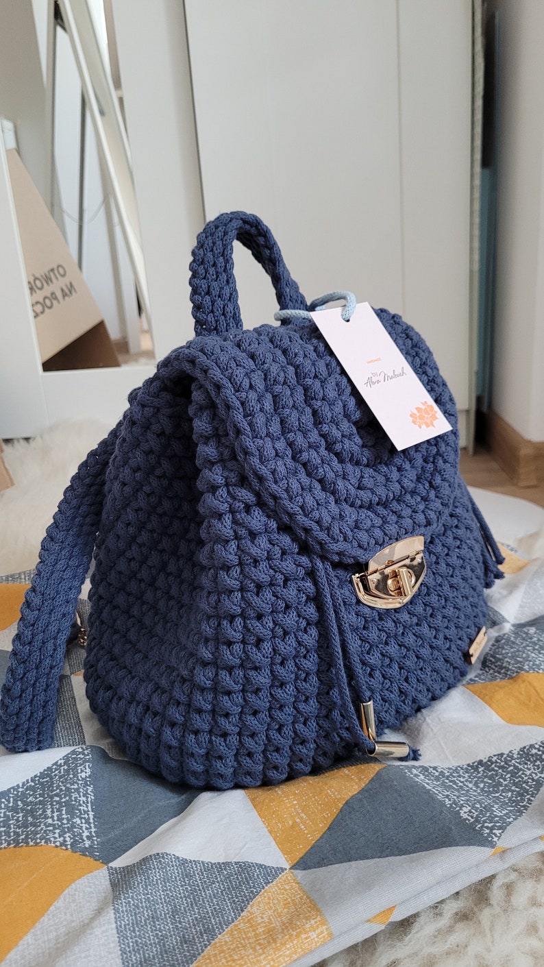 May include: A navy blue crocheted backpack with a gold metal clasp and a small tag that says "by Anna Nadel." The backpack is sitting on a patterned fabric with yellow, grey, and white triangles.