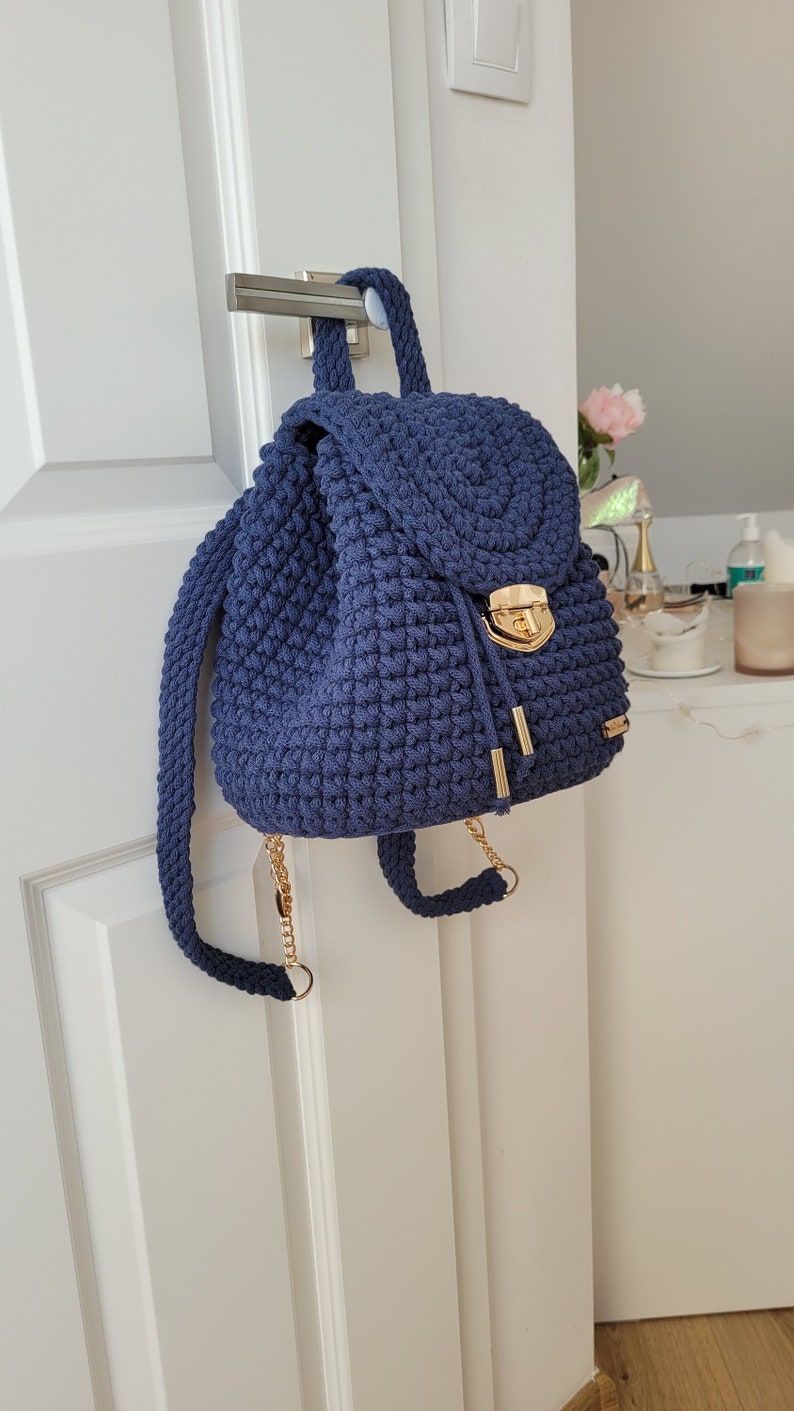 May include: A navy blue crocheted backpack with a gold chain strap and a gold clasp. The backpack is hanging on a white door.