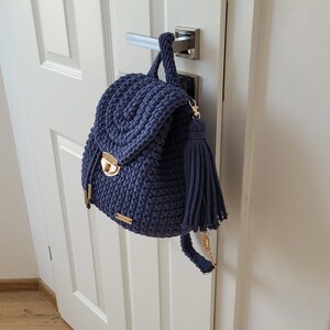 May include: A navy blue crocheted backpack with a gold clasp and a tassel. The backpack is hanging on a white door.