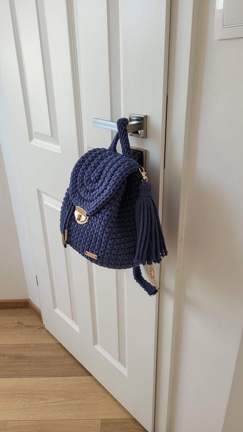 May include: A navy blue crocheted backpack with a gold clasp and a tassel. The backpack is hanging on a white door.