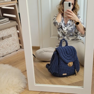 May include: A navy blue crocheted backpack with a gold clasp and a tassel. The backpack is sitting on a wooden floor.
