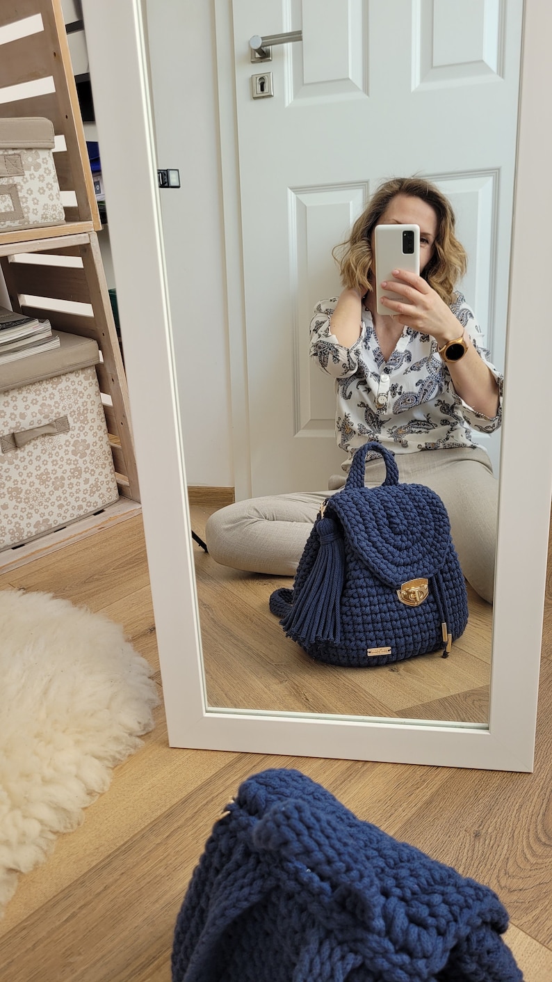 May include: A navy blue crocheted backpack with a gold clasp and a tassel. The backpack is sitting on a wooden floor.