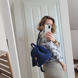 May include: A woman is wearing a white blouse with a floral pattern and beige trousers. She is carrying a dark blue crocheted backpack with a gold zipper and tassel.