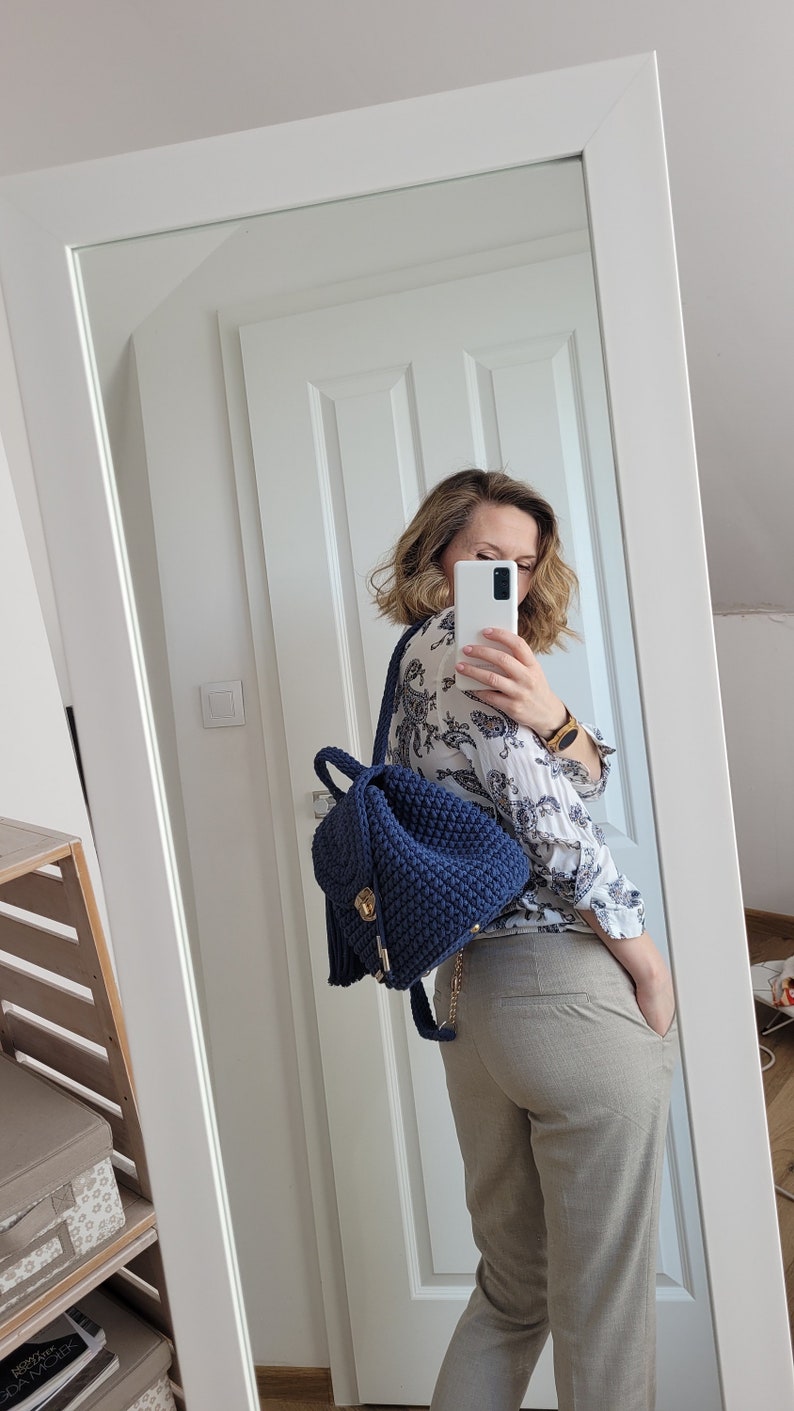 May include: A woman is wearing a white blouse with a floral pattern and beige trousers. She is carrying a dark blue crocheted backpack with a gold zipper and tassel.