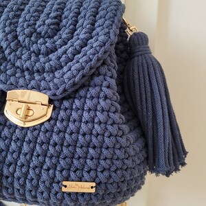 May include: A navy blue crocheted backpack with a gold clasp and a matching tassel. The backpack has adjustable straps and a small gold tag that reads "By Aleksi Malawich".