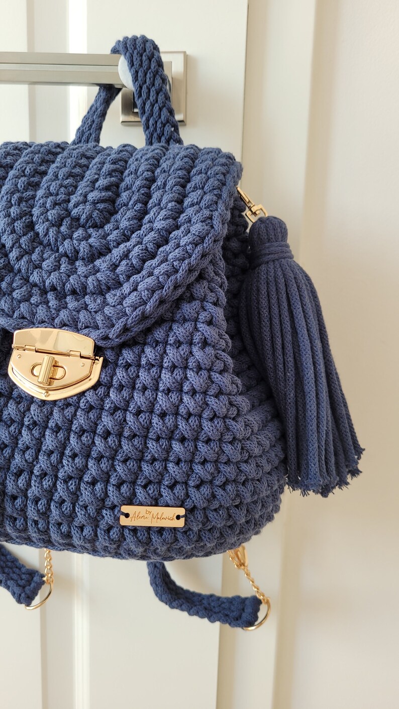 May include: A navy blue crocheted backpack with a gold clasp and a matching tassel. The backpack has adjustable straps and a small gold tag that reads "By Aleksi Malawich".