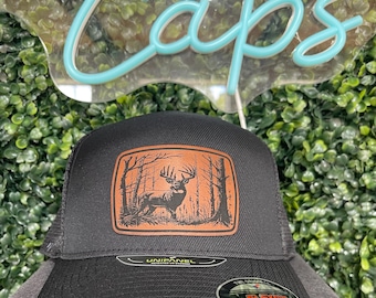 Puede incluir: Gorra de camionero negra con un parche de cuero marrón que presenta la silueta de un ciervo en un bosque. La gorra tiene una parte trasera de malla y una etiqueta Unipanel.