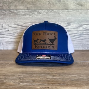 May include: A blue and white trucker hat with a brown leather patch. The patch features the words "Top Notch Kennels" and silhouettes of two dogs and a deer. The hat has a curved brim and an adjustable snap closure.