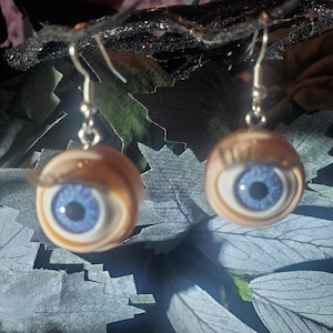 May include: A pair of unique eyeball earrings. Each earring features a round, tan-colored eye with a blue iris and black pupil. The earrings have silver-colored hooks and are set against a backdrop of dark green and gray leaves.