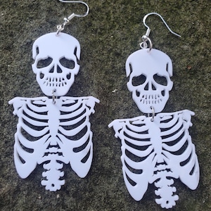 May include: A pair of white skeleton earrings. Each earring features a skull with a ribcage underneath. The earrings are made of plastic and have silver hooks.