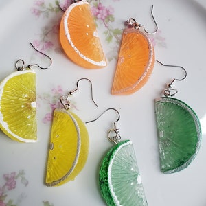 May include: Three pairs of fruit slice earrings. The earrings are made of resin and feature a lime, lemon, and orange slice. The earrings are silver in color and have a hook closure.