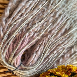 May include: A bundle of synthetic hair extensions in shades of light blonde and pink. The hair is braided and twisted, creating a textured look. The extensions are tied together with a black elastic band.