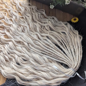 May include: A bundle of wavy, light blonde dreadlocks. The dreadlocks are tied together with a light gray ribbon. The hair is lying on a wooden surface with a black background and a small painting.