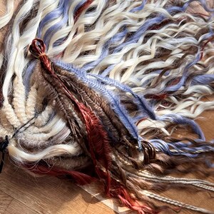 May include: A collection of handmade dreadlocks in various colors, including white, blue, brown, and red. The dreadlocks are arranged on a wooden surface, showcasing their texture and color variations. The image highlights the craftsmanship and the diverse color palette of the hair extensions.