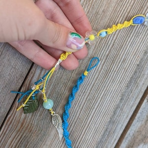 Peut inclure: Un bracelet d'amitié bleu et jaune avec une pierre verte, une pierre bleu clair, un charme d'aile d'ange argenté et un charme de fleur jaune.