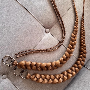Puede incluir: Dos extensiones de cabello castaño claro, largas y gruesas, trenzadas en una sola trenza. Cada trenza está unida a un anillo de metal plateado en la parte superior. Dos mechones más finos y sin trenzar sobresalen de los anillos.
