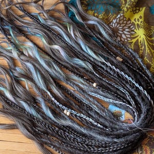 Könnte beinhalten: Eine Sammlung grauer, schwarzer und blauer Kunsthaar-Dreadlocks. Einige Dreadlocks sind geflochten, andere sind locker und wellig. Holzperlen sind eingestreut. Die Dreadlocks liegen auf einer Holzoberfläche mit gemustertem Stoff im Hintergrund.