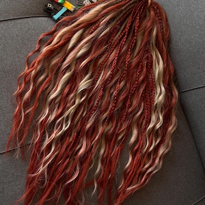 May include: A set of synthetic hair extensions featuring a blend of wavy and braided strands in shades of deep red, copper, and light blonde. The hair is gathered at the top with a small elastic band, and a collection of colorful plastic building bricks.