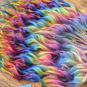 Puede incluir: Un conjunto de rastas de colores del arcoíris, con una mezcla de tonos vibrantes como rojo, amarillo, azul y morado. El cabello sintético tiene una textura ondulada y está atado con una cinta marrón. Las rastas están dispuestas sobre una superficie de madera.