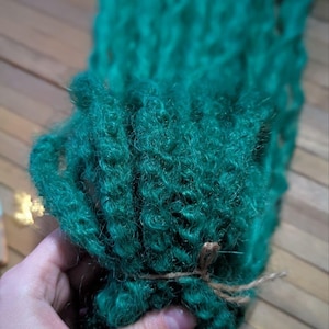 May include: A bundle of teal green synthetic dreadlocks tied together with twine. The dreadlocks are made of soft, curly fibers.