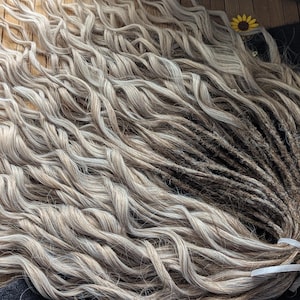 Puede incluir: Un conjunto de rastas onduladas de color gris claro con raíces más oscuras, atadas con una cinta blanca. Las rastas tienen una apariencia natural y texturizada, lo que sugiere un accesorio para el cabello hecho a mano o personalizado.