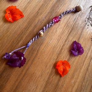 Peut inclure: Une baguette artisanale avec un cordon lavande, des perles en bois et un cristal rose. La baguette est ornée de deux breloques en forme de feuilles violettes et oranges. Les objets sont disposés sur une surface en bois.