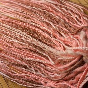 May include: A bundle of pink and brown synthetic dreadlocks. The dreadlocks are a mix of light pink, peach, and brown tones, with varying textures and braiding styles. The hair is held together at one end.