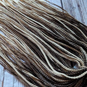 Könnte beinhalten: Ein Bündel vorgeflochtener Haarverlängerungen in Braun-, Blond- und Hellblondtönen. Das Haar ist fächerförmig angeordnet, die Enden der Zöpfe sind zusammengefasst.