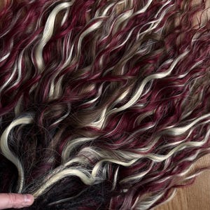 May include: A bundle of wavy hair extensions in shades of burgundy, cream, and brown. The hair is arranged in a flowing pattern, with the colors intermingling to create a textured appearance. The extensions are likely for styling or adding volume.